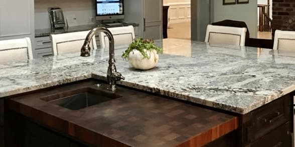 Granite kitchen island with a butcher-block section around the sink and white chairs